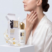 A young woman in a white silk robe applies a drop of serum to her neck, seen looking sideways. The 'mastic spa' Instant Lifting Serum (Donkey Milk & Mastic Oil) bottle and box are on a white plinth, accompanied by an artistic splash of dense white donkey milk and embedded gold mastic resin droplets.