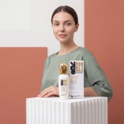 A young woman in a sage green top stands confidently behind a fluted white geometric plinth, her hand resting near the 'mastic spa' Instant Lifting Serum bottle and box. The background features modern white and terracotta panel art.
