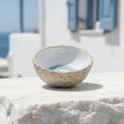 Hand-thrown ceramic bowl resting on a white marble surface with a blurred Greek village background.