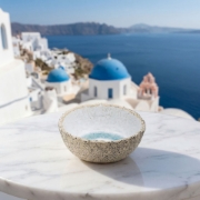 A textured ceramic bowl on a marble table overlooking the blue domes and Aegean Sea in Santorini, Greece.