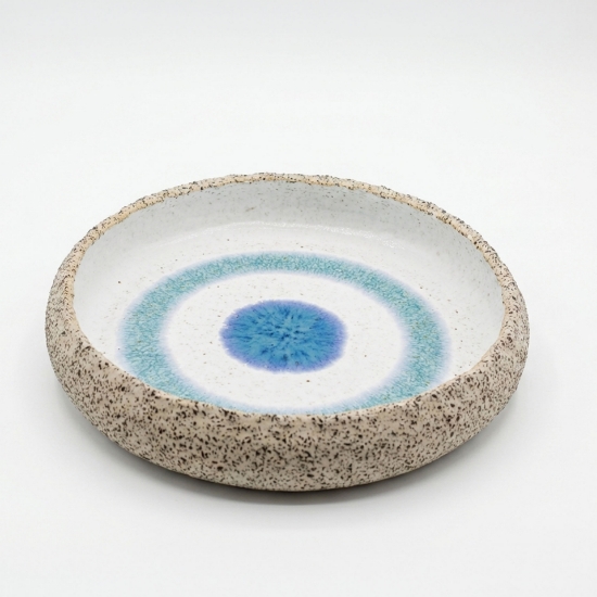 Handmade ceramic bowl with a speckled exterior and a blue "evil eye" concentric circle design on a white background.