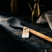 An olive wood spatula with a black silicone head resting on a linen cloth next to a small ceramic bowl of flour and rosemary, with sunlit potted herbs in the background.