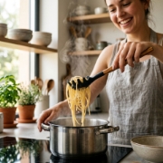A woman smiling while using a wooden-handled spaghetti spoon to lift pasta from a steaming pot.