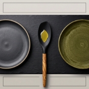 Minimalist editorial flat lay of a silicone spoon with olive oil, flanked by ceramic plates on a slate background.