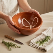 Hands holding a large hand-thrown terracotta salad bowl with a white loop design, on a counter with herbs and a knife