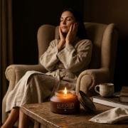 Aurea Gold Scented Candle lit on wooden table, woman relaxing in linen robe
